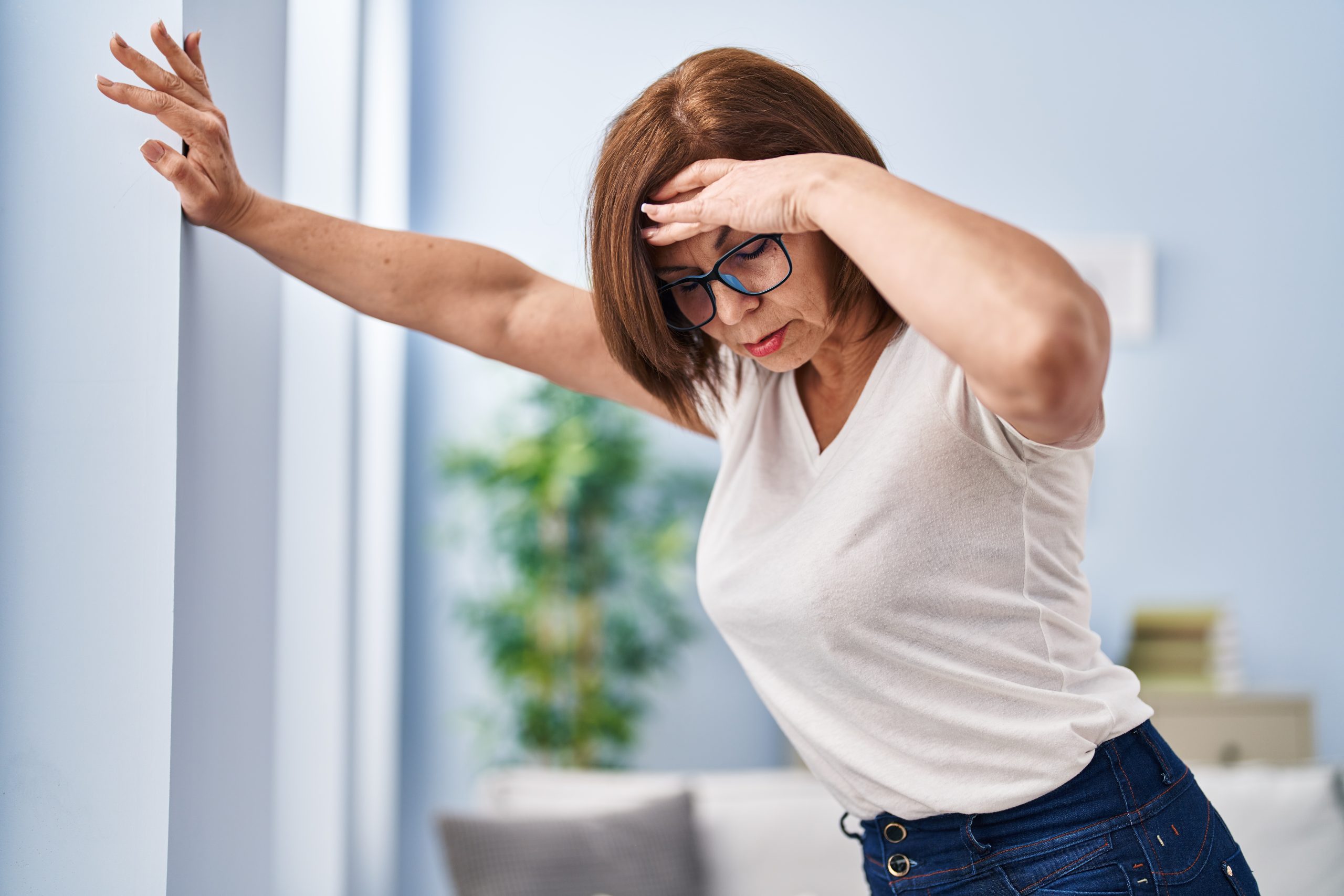 Woman leaning against a wall with one hand on her forehead, appearing unsteady and dizzy in a home setting.